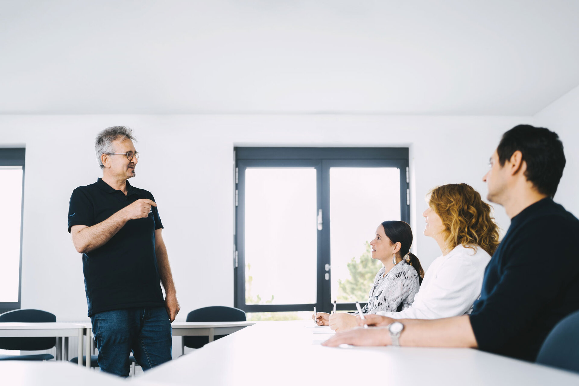 Es findet ein Beratungsgespräch statt. Ein Ingenieure steht vor zwei Kunden, die vor ihm sitzen und präsentiert die individuellen Lösungsvorschläge zur Baugruppenreinigung. 
   | © @The Sour Cherry Fotografie - Michaela Curtis